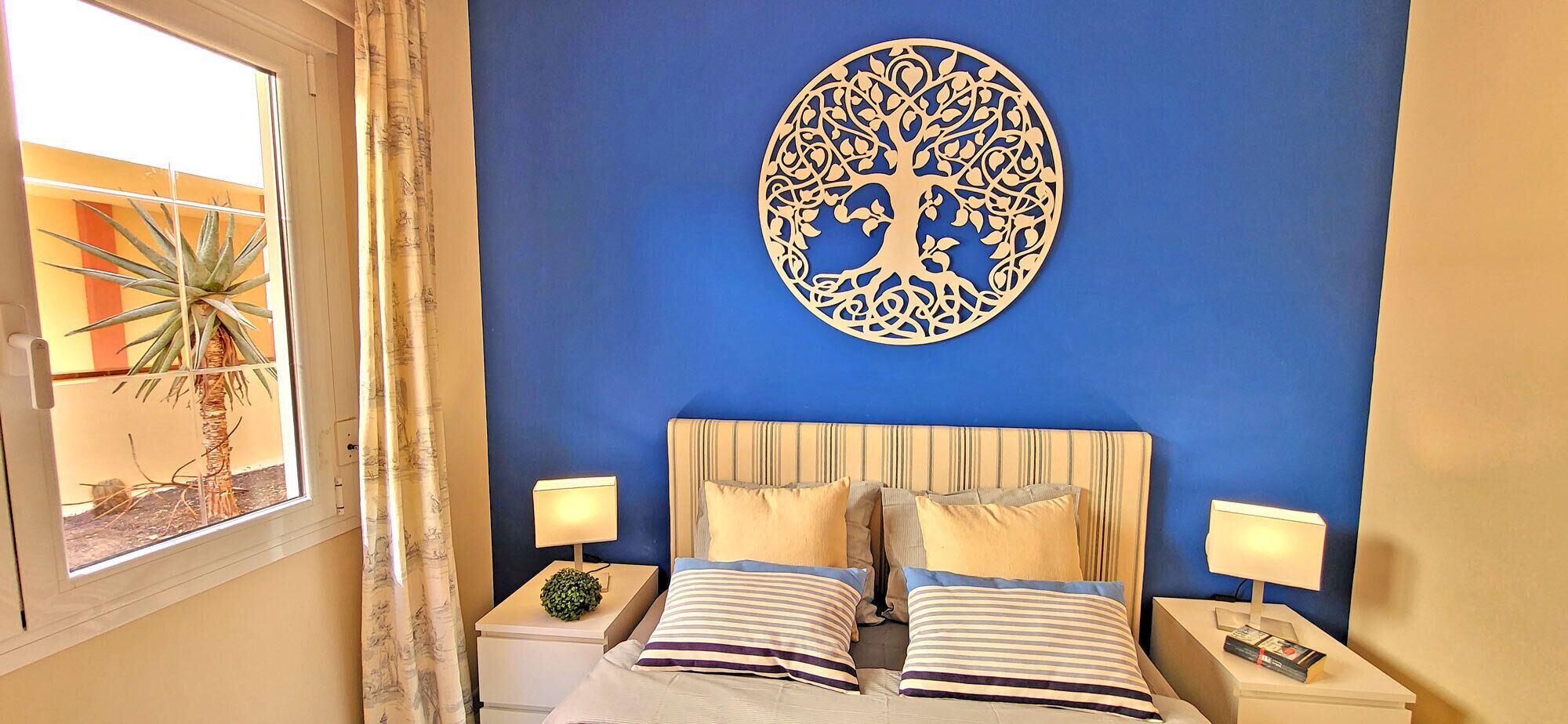 A bedroom with a bed, two bedside tables, a wall-mounted decorative piece, and a window with curtains.