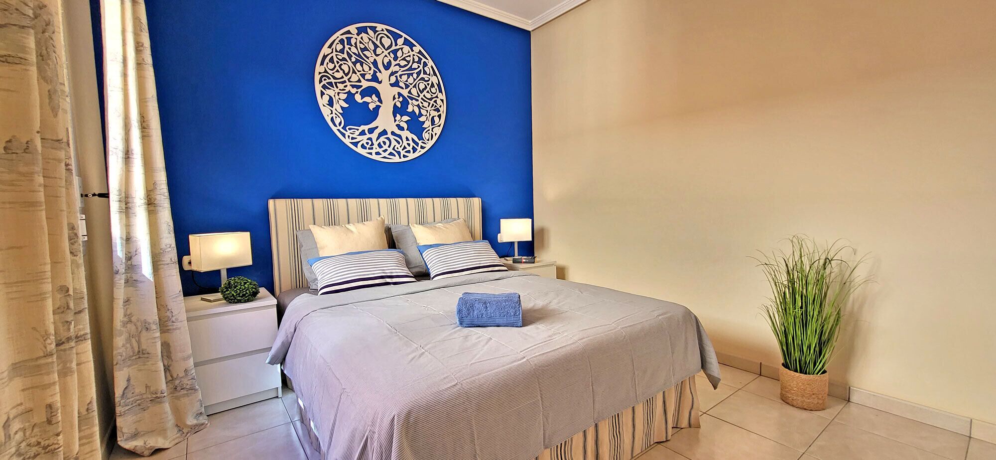 A bedroom with a bed, nightstands, a blue wall with a decorative tree, and a potted plant.