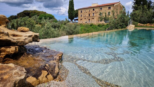 Una piscina cristalina con borde de piedra, un edificio de piedra con amplias ventanas, y vegetación circundante.