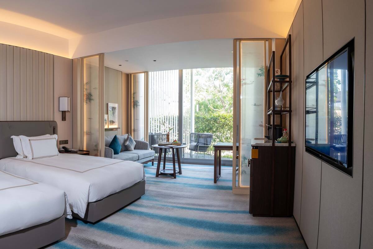Habitación de hotel con dos camas, un sofá, un balcón con sillas y un televisor.