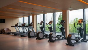 A gym with treadmills, elliptical machines, and stationary bikes arranged in a row with large windows offering a view of greenery. - Nanhu Hotel (Jiaxing)