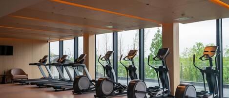 Ett gym med löpband, crosstrainers och motionscyklar uppställda i en rad, med stora fönster som ger utsikt över grönområden.