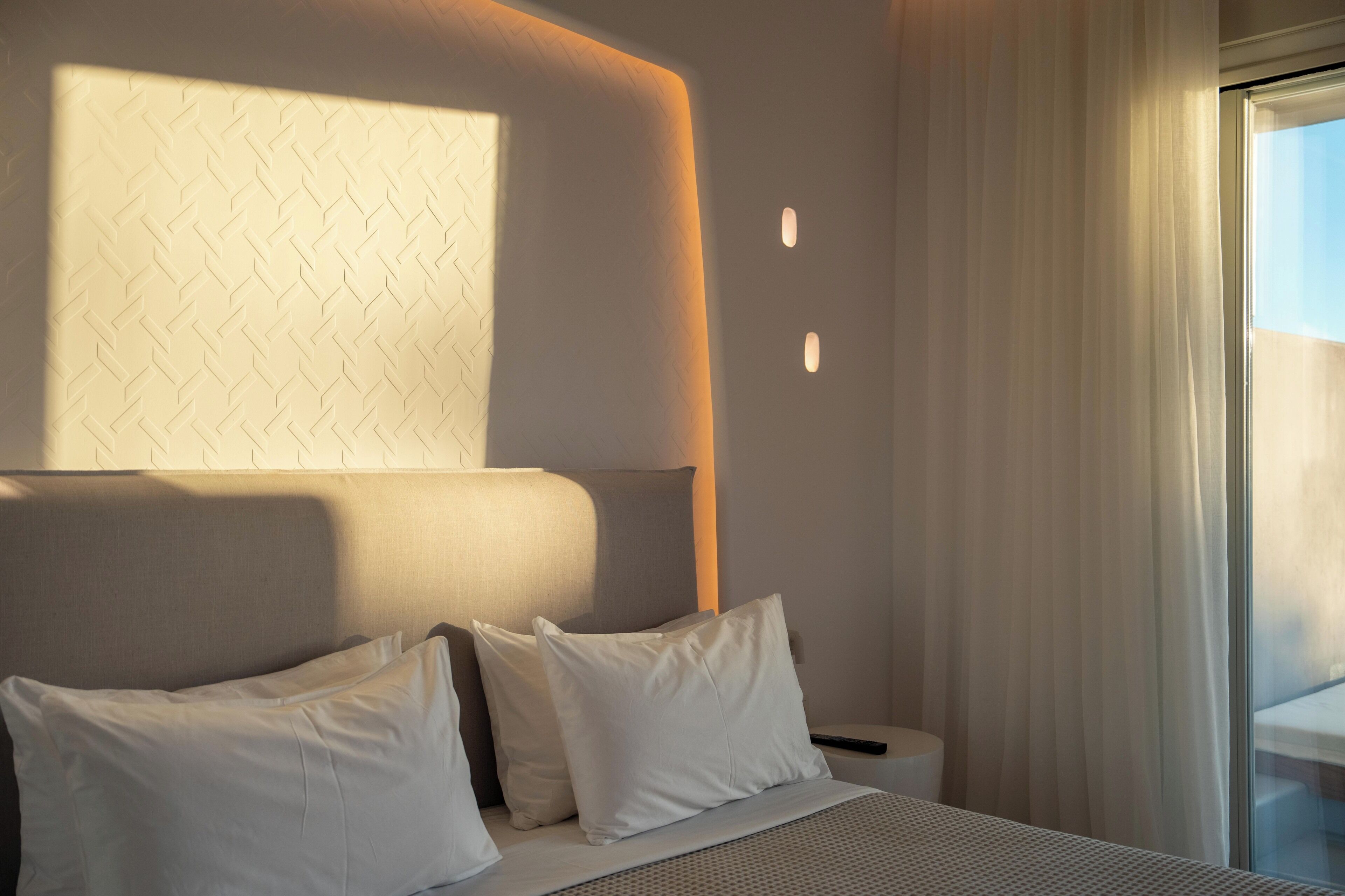 Una habitación de hotel moderna con una cama grande, ropa de cama blanca, un cabecero con textura y una mesita de noche con un control remoto, además de una ventana con cortinas translúcidas.