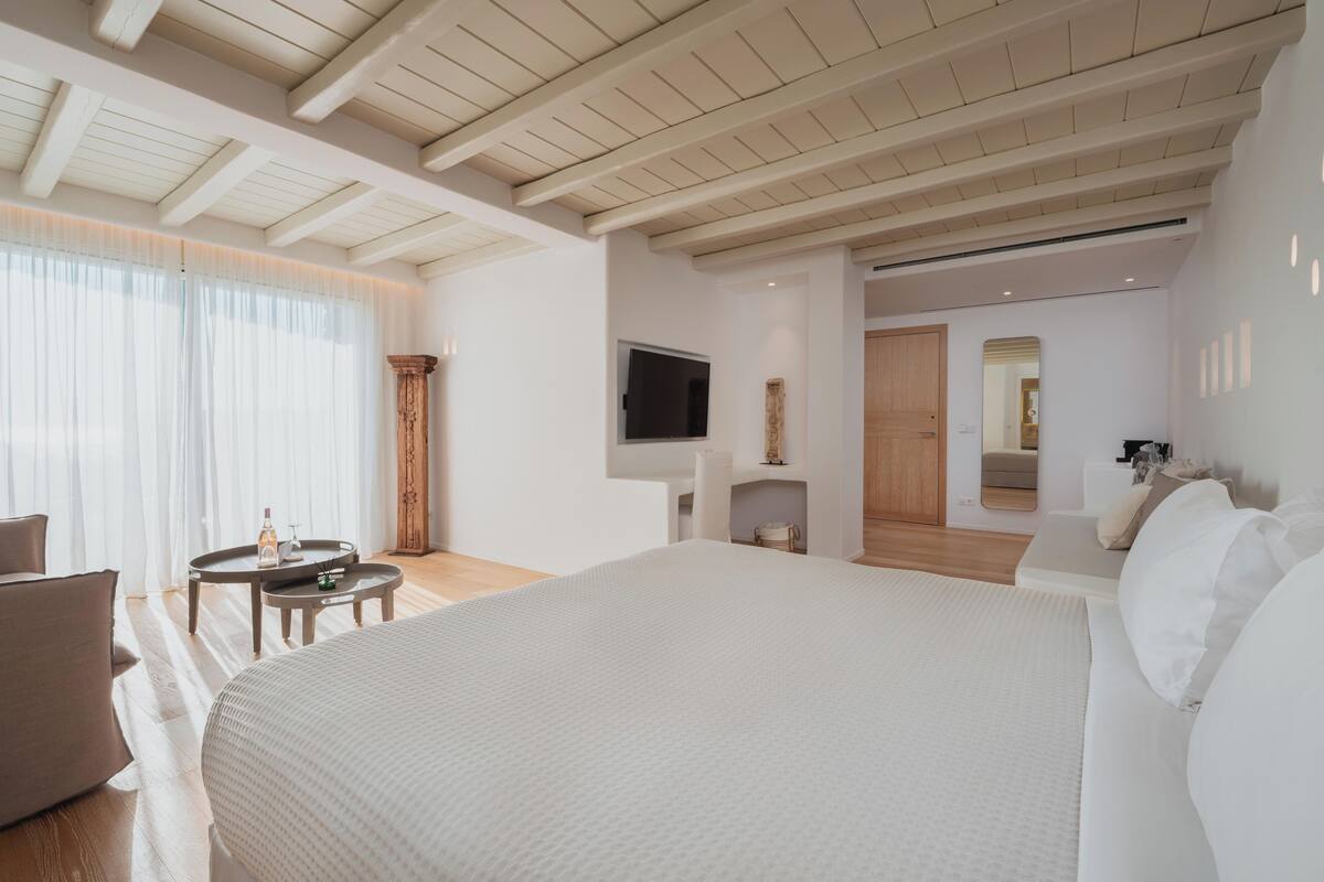 Une chambre d’hôtel moderne avec un grand lit, un téléviseur fixé au mur et une petite table sur laquelle se trouvent une bouteille et des verres.