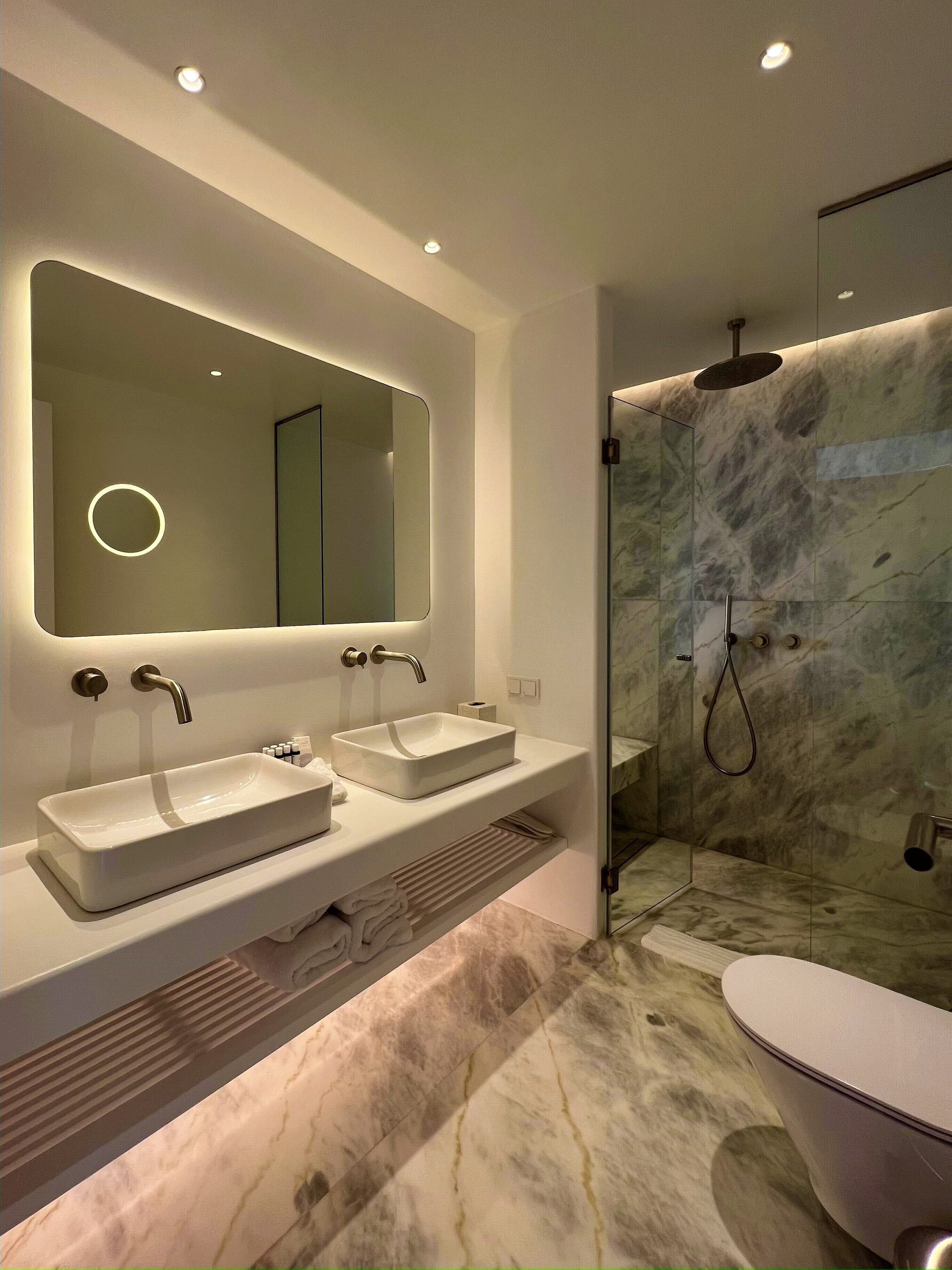 A modern bathroom with two sinks, a large mirror, and a shower area with marble walls.
