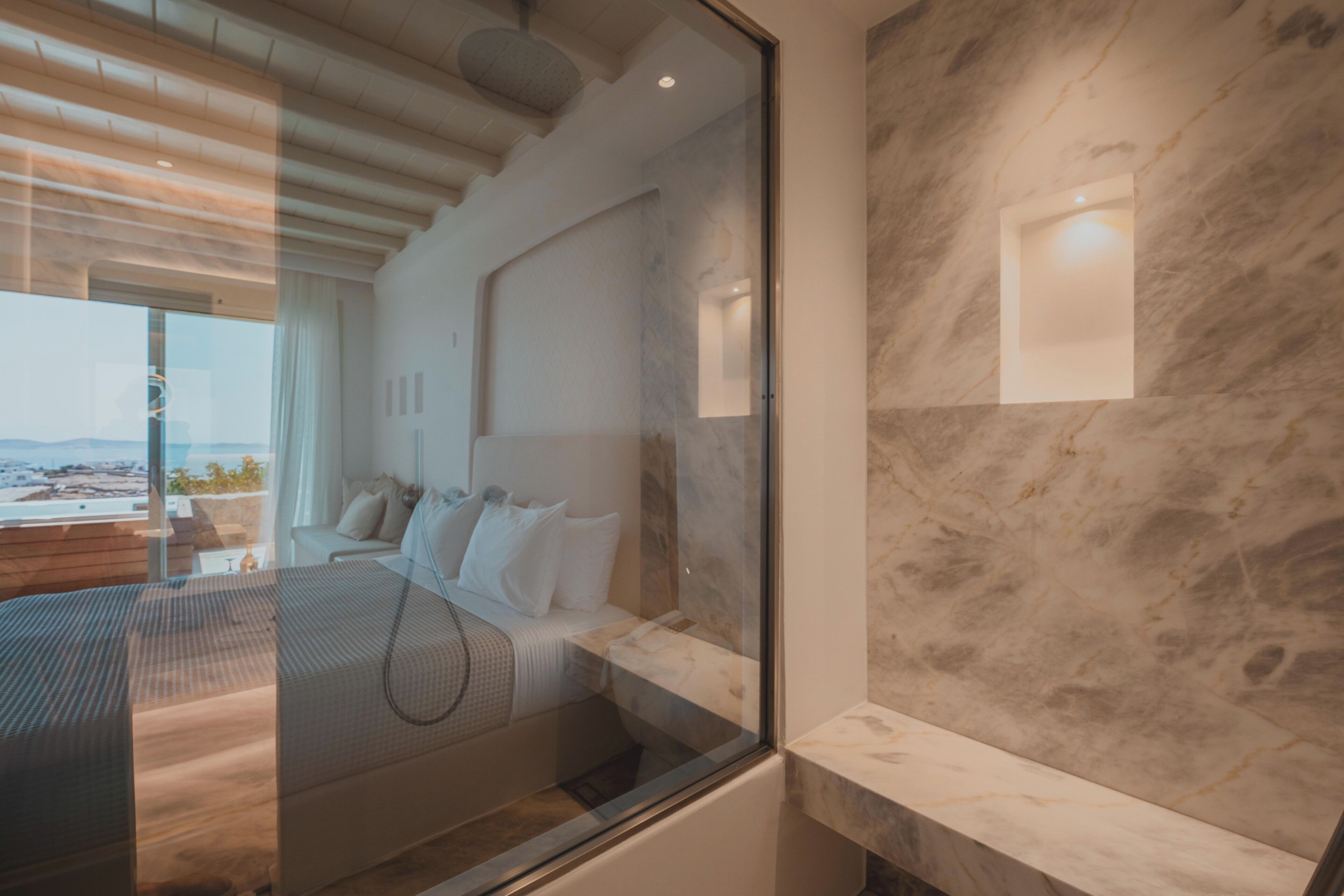 A modern hotel room with a large bed, a glass partition, and a view of the city.