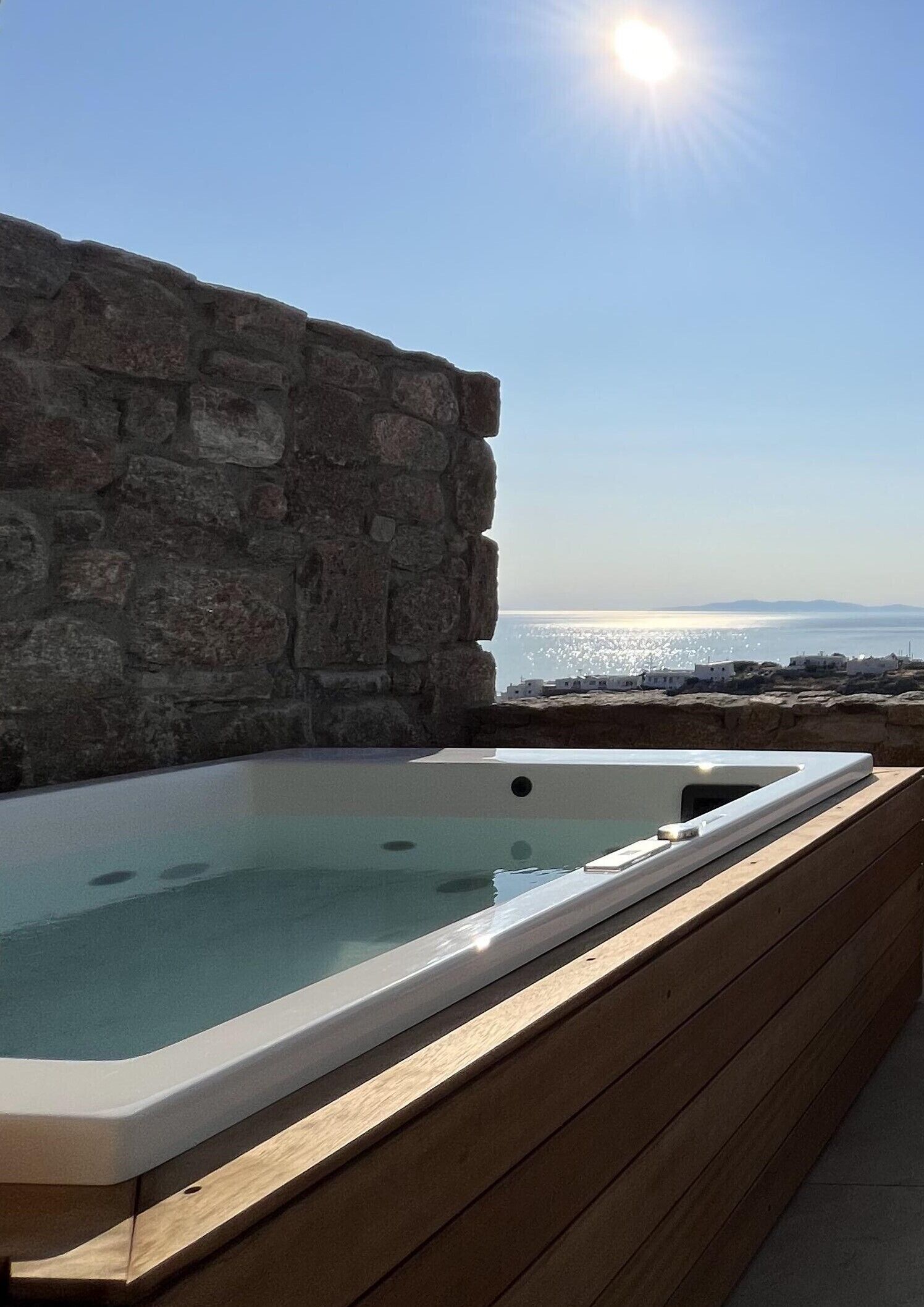 Muro de piedra con terraza de madera y bañera de hidromasaje con vista a un cuerpo de agua.