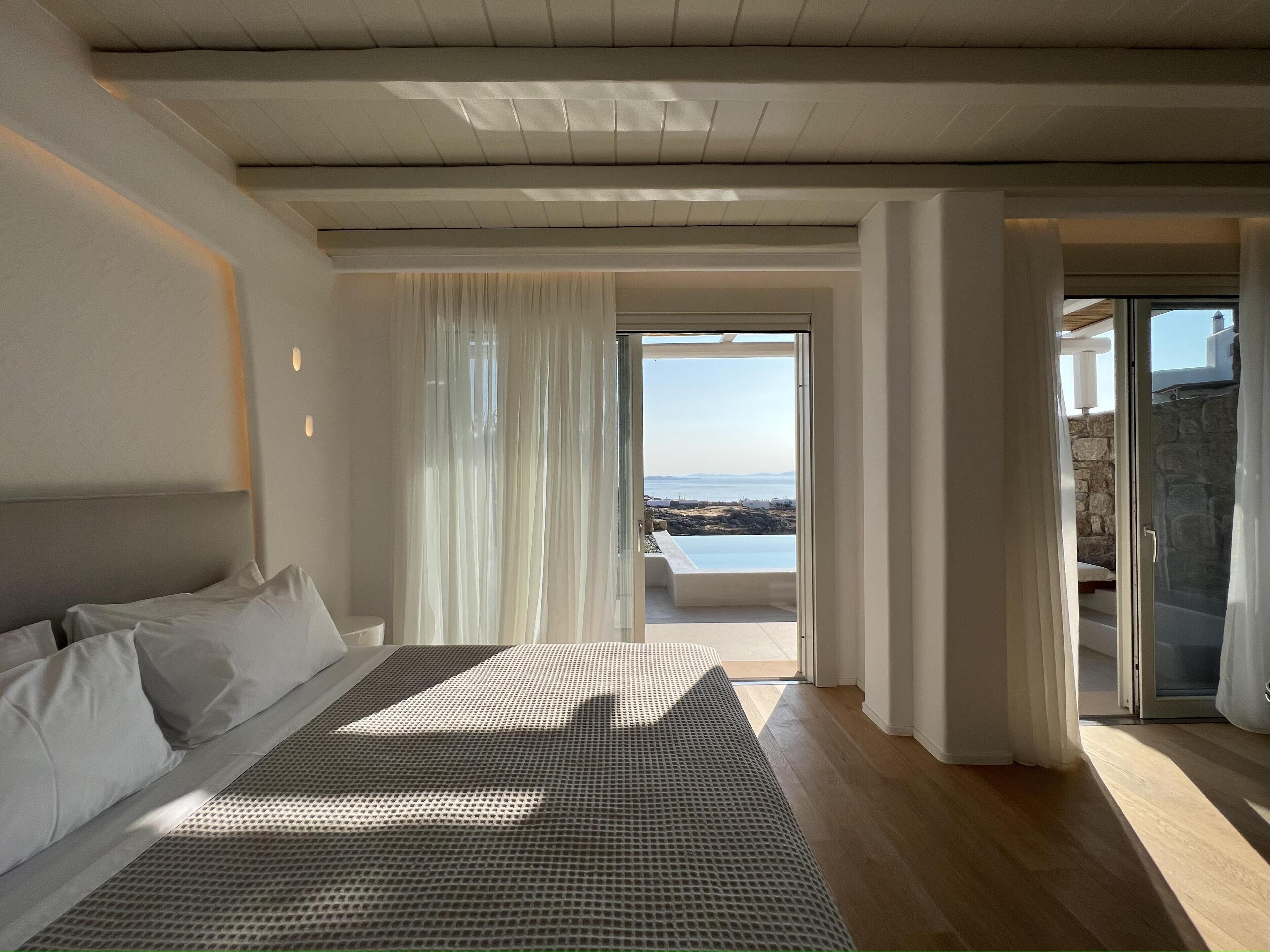 A bedroom with a large bed, a view of the sea through a glass door, and a stone wall.