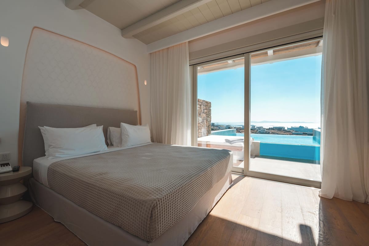 Une chambre avec un grand lit, une table de chevet et une vue sur une piscine et des immeubles, visible à travers une porte-fenêtre coulissante.