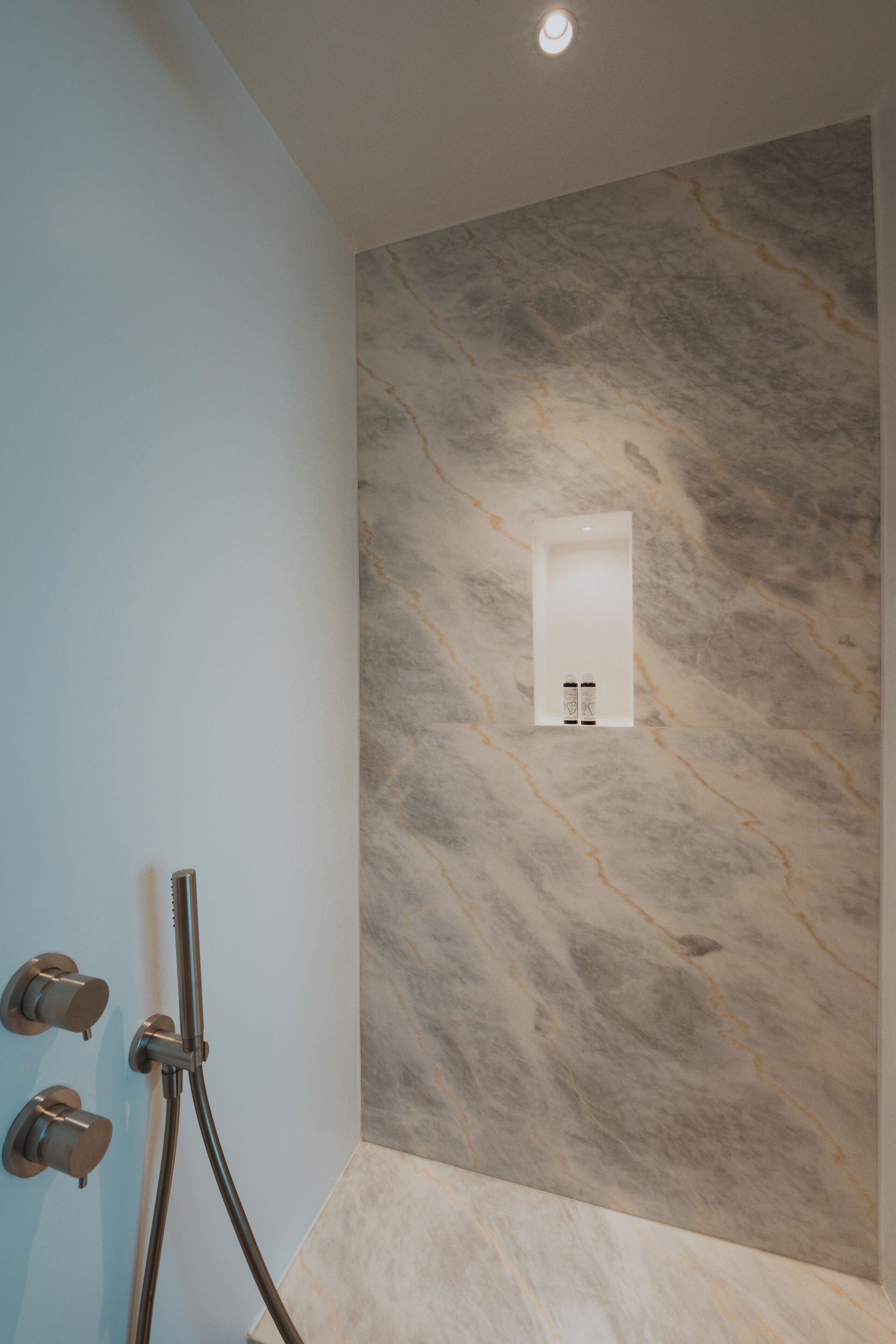 A bathroom with a marble wall, a shower with a handheld showerhead, and a wall-mounted light fixture.