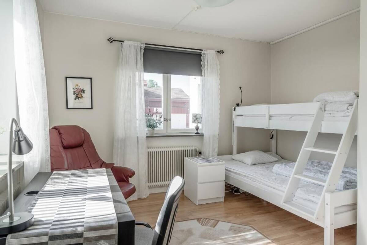 A room with a bunk bed, a desk with a lamp, a chair, a radiator, and a window with curtains.
