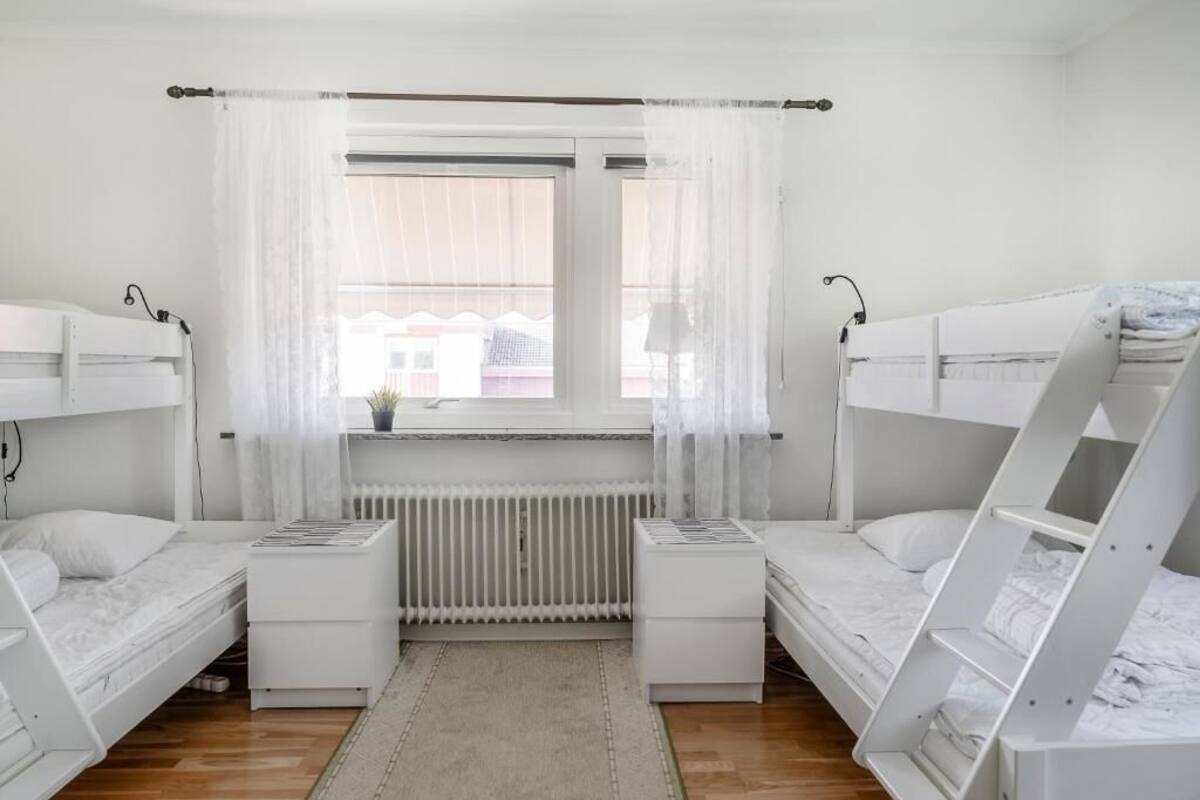 A room with two bunk beds, a radiator, a window with sheer curtains, and a small table with a lamp.