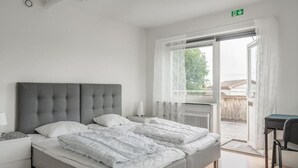 A bedroom with a grey upholstered bed, white bedding, a bedside table with a lamp, and a view of the outside through a glass door. - Vandrarhemmet Stranden i Mora (Mora)