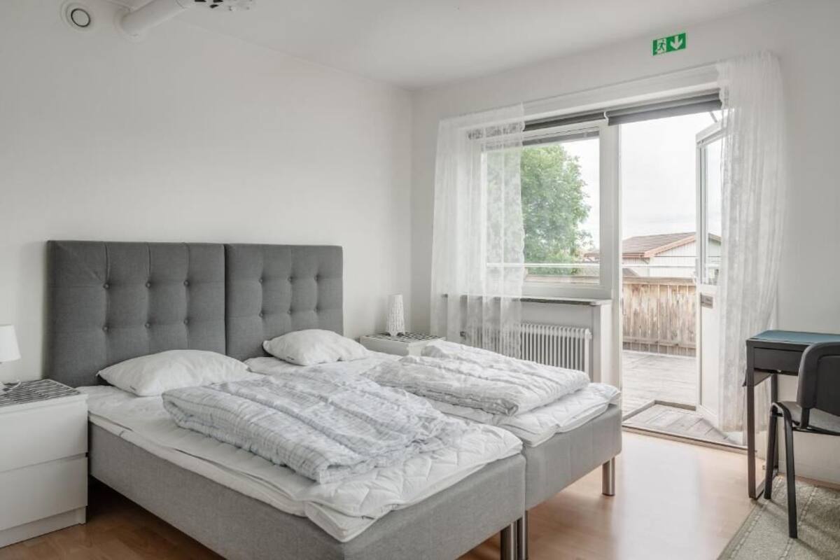 A bedroom with a grey upholstered bed, white bedding, a bedside table with a lamp, and a view of the outside through a glass door.