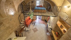A stone-walled room with a spiral staircase, a sofa, a small table, and a bed in the background. - PA - Pandora Loft  (Valletta)