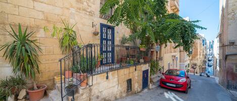 Une rue étroite où est garée une voiture rouge sur le bord, avec des plantes en pot et un immeuble doté d’une porte bleue et d’une rambarde en fer.