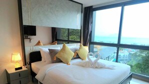 Habitación de hotel con una cama grande, una mesita de noche con lámpara y vistas de la ciudad a través de amplias ventanas.