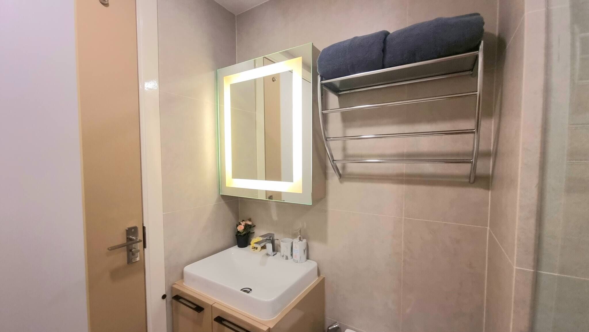 Une salle de bain avec un lavabo blanc, un miroir avec éclairage indirect, un porte-serviettes et une porte.