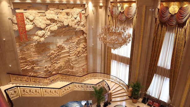 Um amplo hall de hotel com uma escadaria de mármore, detalhes esculpidos nas paredes e um grande lustre.