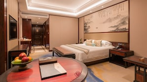 Quarto de hotel com cama, mesas de cabeceira, escrivaninha, cadeira, cesta de frutas e parede com caligrafia.