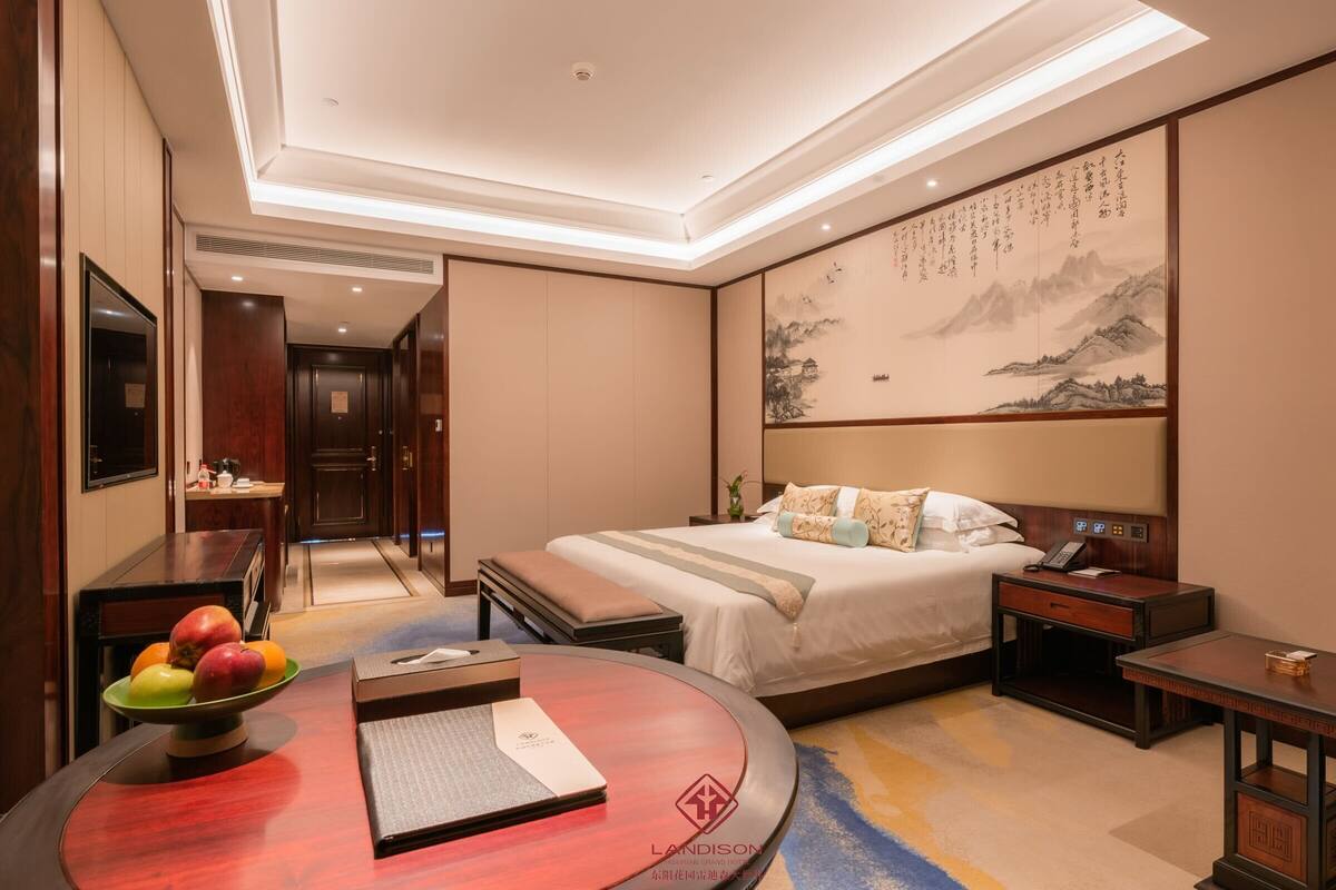 A hotel room with a bed, bedside tables, a desk, a chair, a fruit bowl, and a wall with calligraphy.
