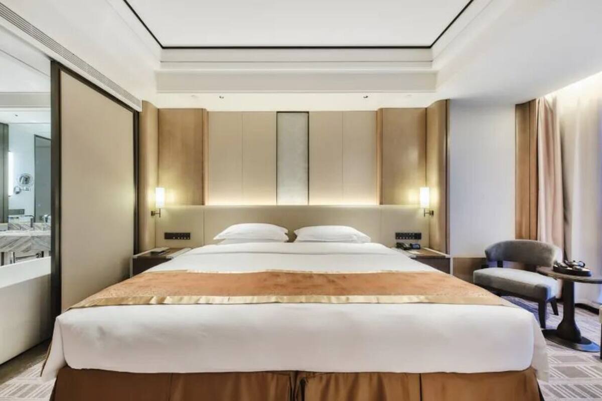 Quarto de hotel moderno com uma cama grande, área de estar com poltrona e mesa, toucador com espelho e uma janela ampla com cortinas.