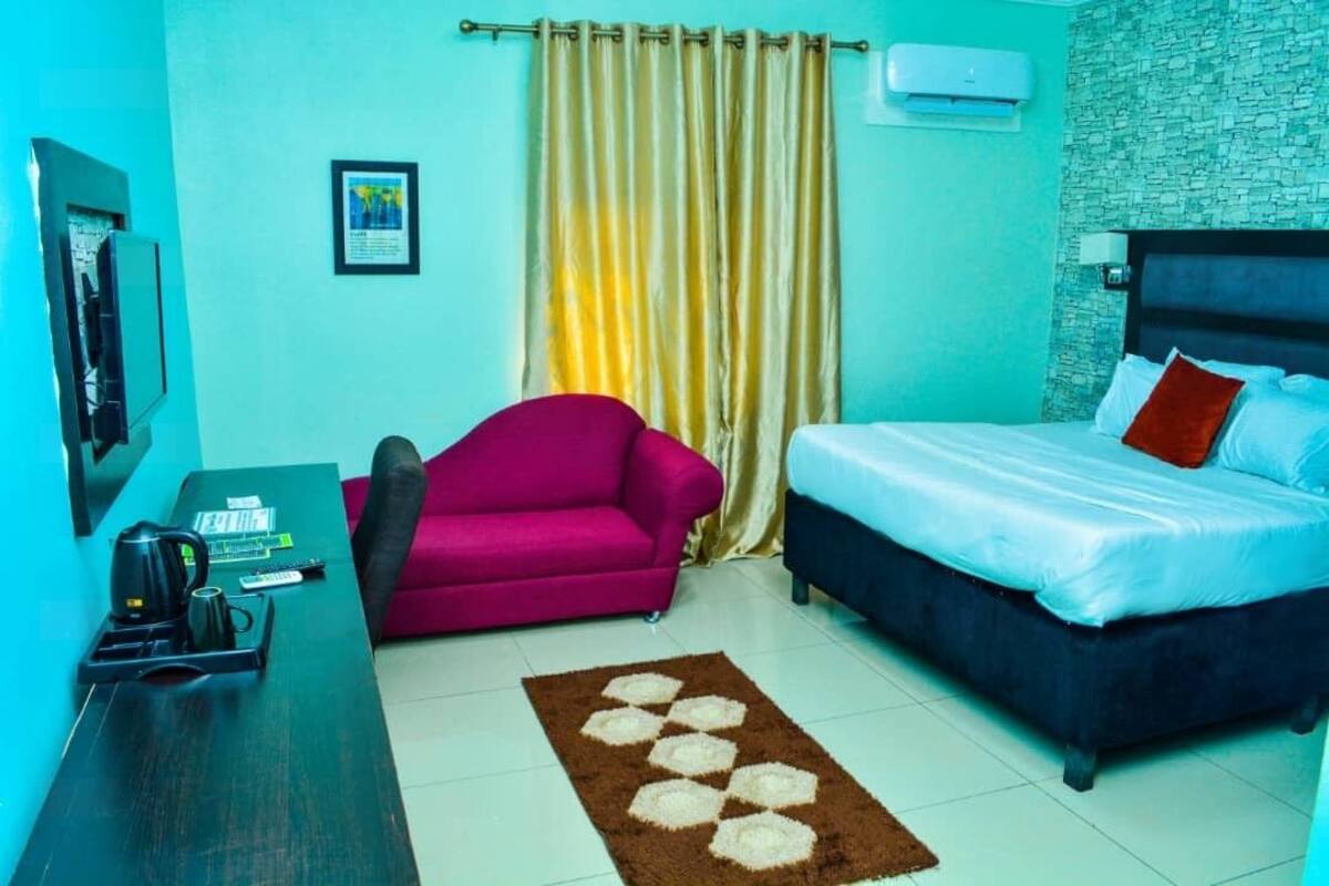 A hotel room with a bed, a desk with a coffee maker, a pink chair, and a curtain.
