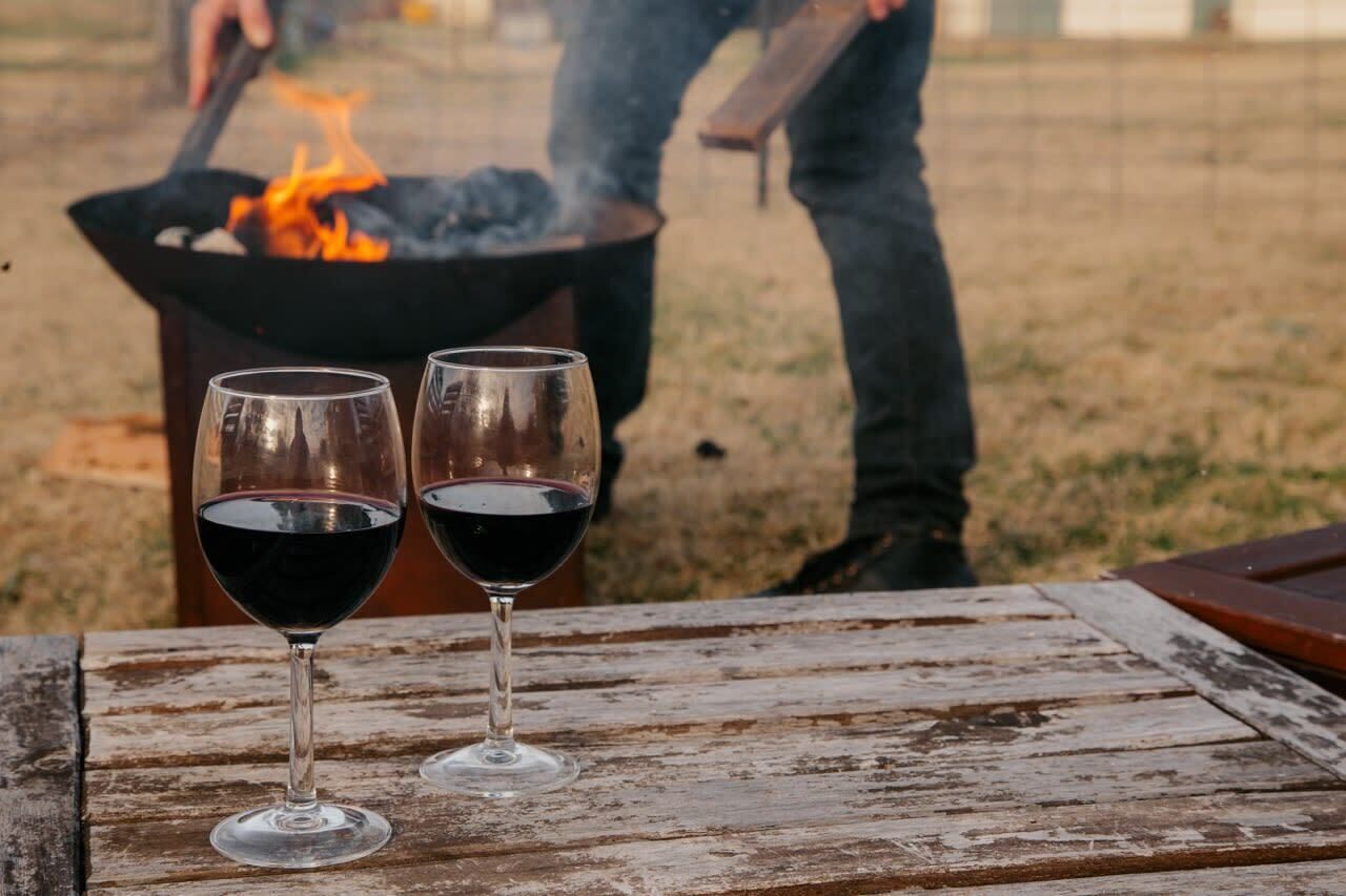 Two wine glasses on a rustic wooden table with a fire pit in the background.