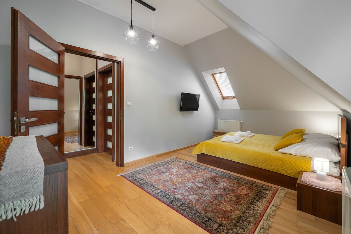 A bedroom with a sloped ceiling, a bed with yellow bedding, a wooden wardrobe, a television on the wall, and a patterned rug.