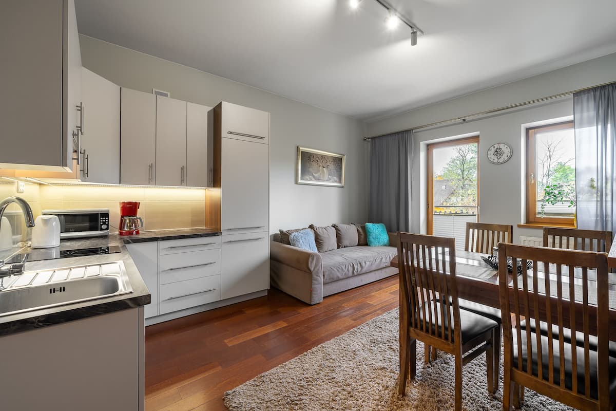 A modern kitchen with white cabinets, a microwave, and a coffee maker. There is a dining area with wooden chairs and a table, and a sofa with cushions.