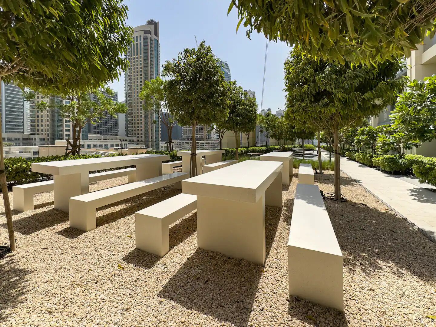 An outdoor seating area with benches and tables, surrounded by trees and a cityscape in the background.