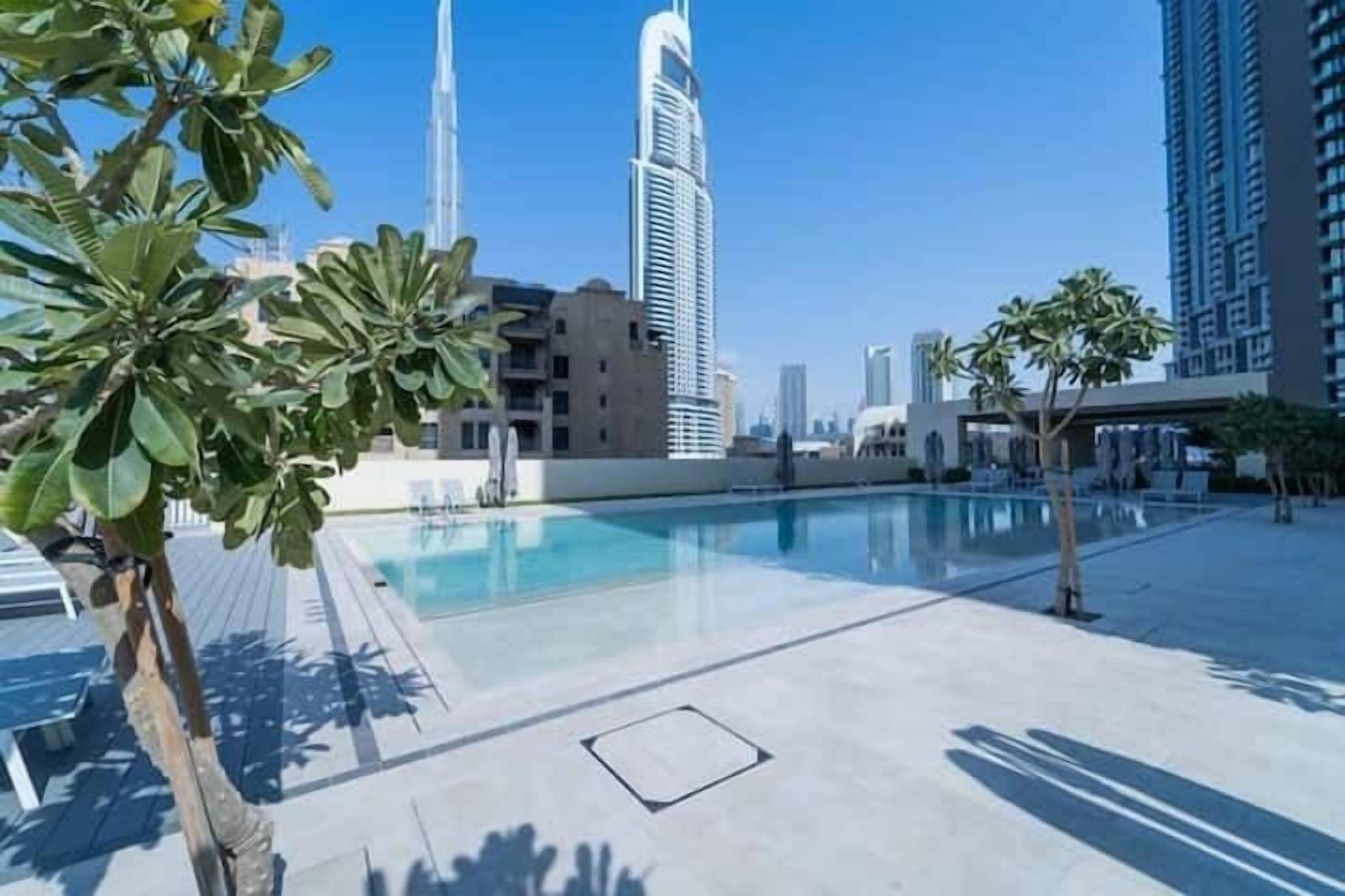 Uma área de piscina no terraço com palmeiras, céu claro e edifícios modernos ao fundo.