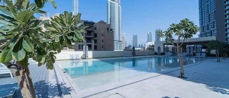 Uma área de piscina no terraço com palmeiras, céu claro e edifícios modernos ao fundo.