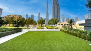 A landscaped area with a lawn and trees, surrounded by modern buildings and a prominent skyscraper in the background. - Vogue| in Heart of Downtown with Burj Khalifa view (Dubai)