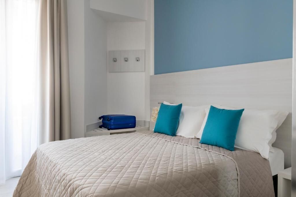 Une chambre d’hôtel avec un lit, une table de chevet, une valise bleue et un mur muni d’un interrupteur.
