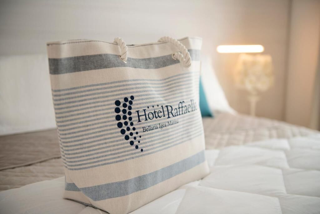 A striped tote bag with the text "Hotel Rafface" and "Bellaria Igea Marina" on a bed.