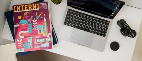Uma mesa com um laptop, uma revista intitulada “INTERNATIONAL DESIGN LANDSCAPES” e duas pequenas plantas em vasos.