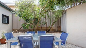 A patio with a glass table and blue chairs, surrounded by a garden and a wooden fence.