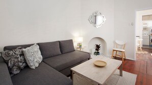 A modern living room with a grey sectional sofa, a wooden coffee table, and a white armchair. A decorative mirror hangs on the wall, and a small table with a lamp is visible.