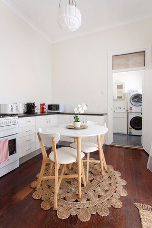 Une cuisine moderne avec des meubles blancs, une table de salle à manger ronde entourée de chaises, un luminaire suspendu et un lave-linge en arrière-plan.
