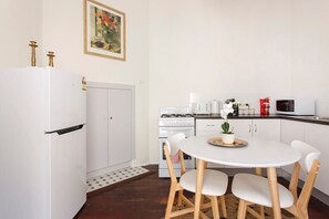 Une cuisine compacte avec des meubles blancs, un réfrigérateur blanc, une cuisinière et un four à micro-ondes. On y trouve une petite table à manger avec quatre chaises.