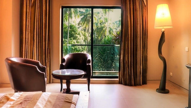 Une chambre d’hôtel avec un lit, deux fauteuils, une petite table, une lampe et une vue sur les palmiers par une grande fenêtre.