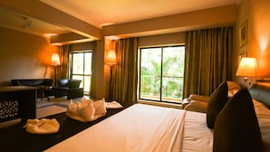 Une chambre d’hôtel avec un grand lit, deux fauteuils, un bureau et une vue sur la verdure par les fenêtres.