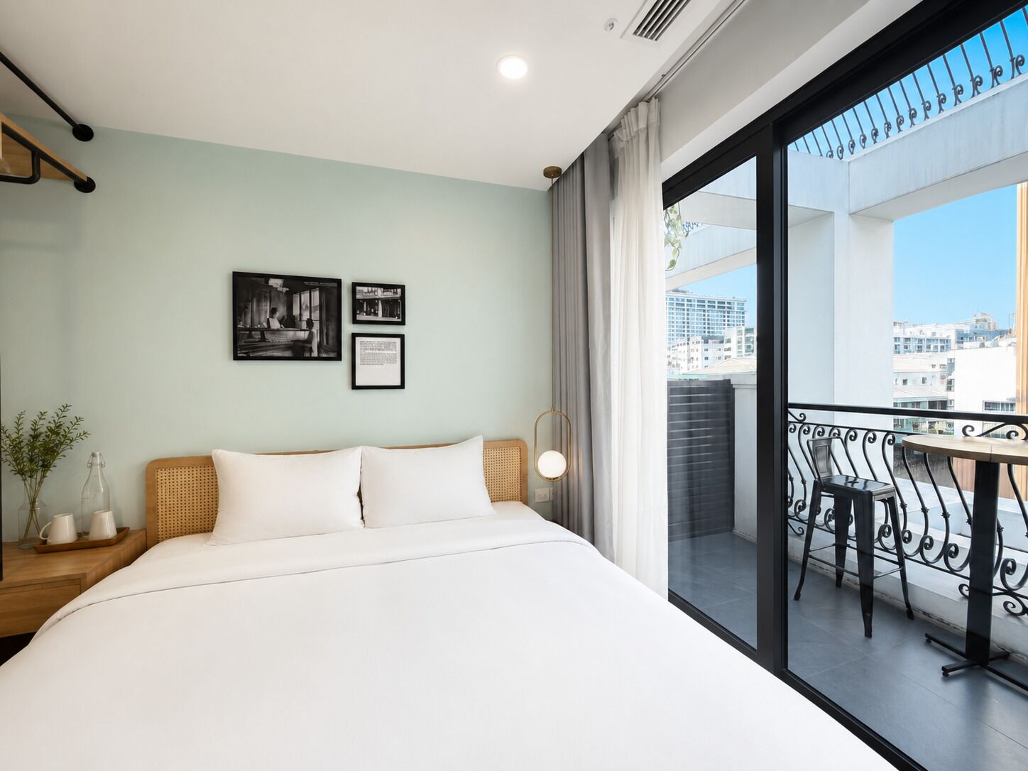 A hotel room with a bed, a small table, a chair, a balcony with a view of buildings, and two framed pictures on the wall.
