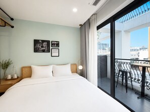 A hotel room with a bed, a small table, a chair, a balcony with a view of buildings, and two framed pictures on the wall. - Wilque Hotel Hanoi (Hanoi)