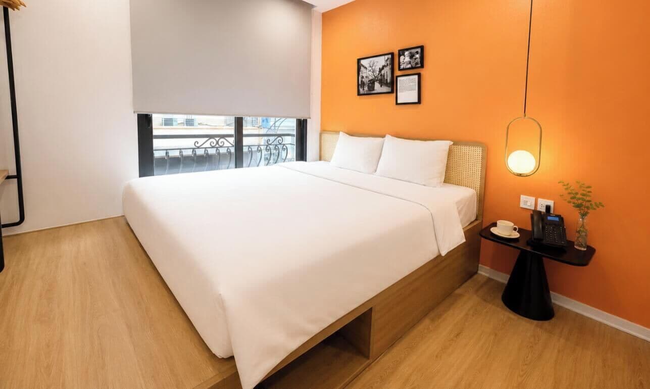 A modern hotel room with a large bed, a bedside table with a lamp, and a view of the outdoors.