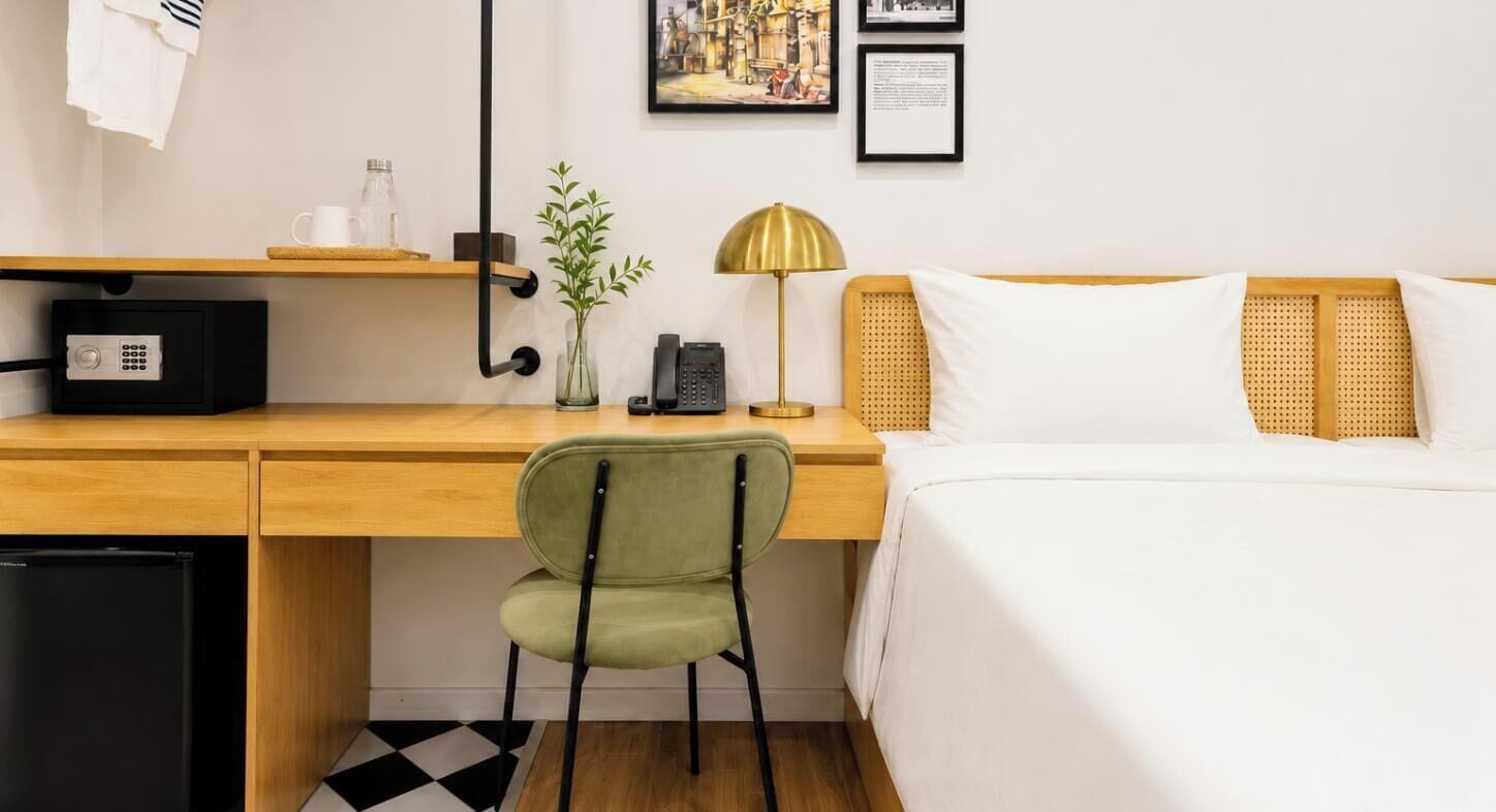 A hotel room with a wooden desk, a green chair, a bed with white bedding, a black refrigerator, a gold lamp, a vase with green plants, and a wall-mounted phone.