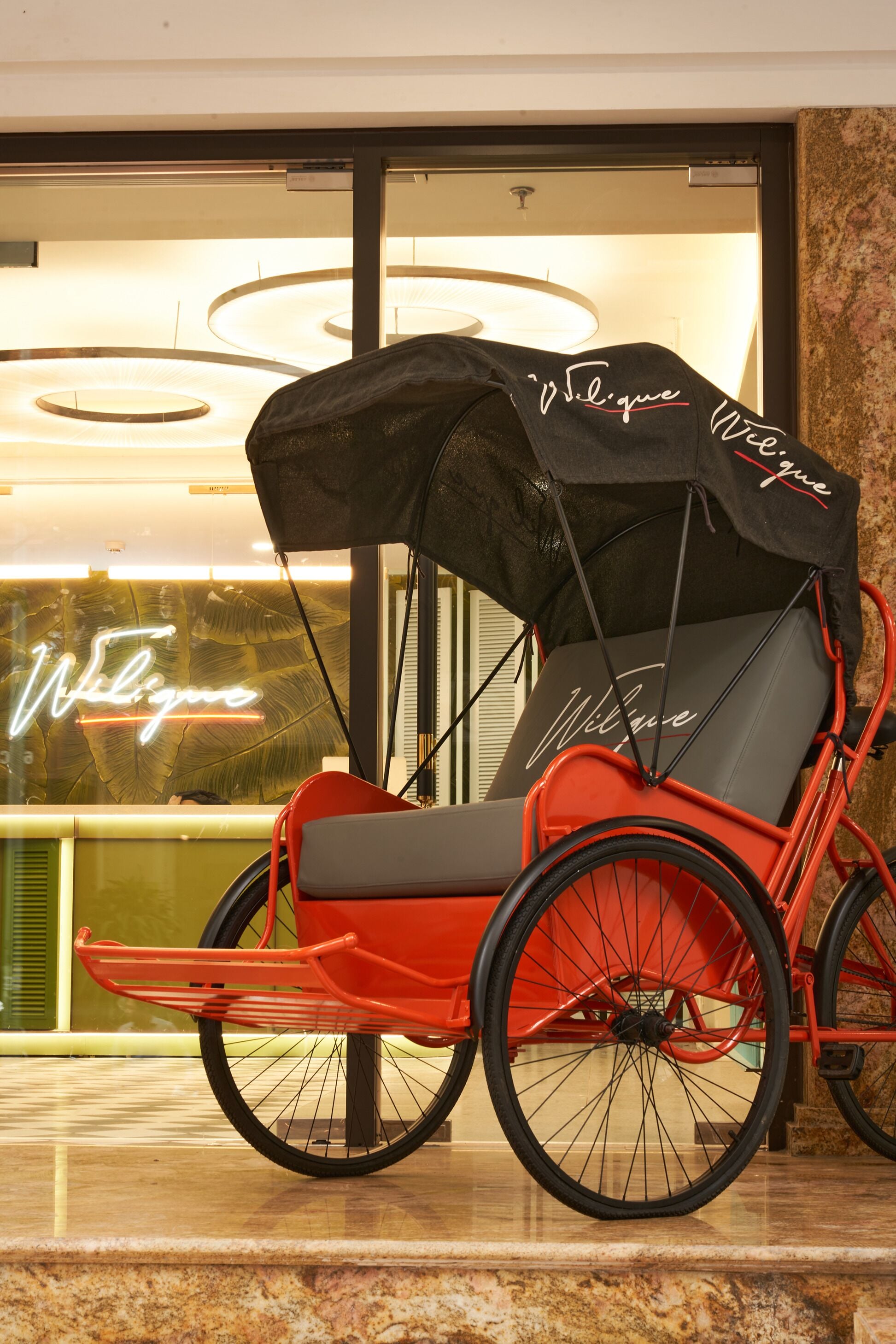 Un fauteuil roulant rouge, doté d’un auvent noir et arborant la marque « Wiligene », est placé devant une porte vitrée surmontée d’une enseigne au néon.