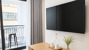 A modern room with a wooden desk, a mounted TV, a chair, a small table with a vase of plants, and a view of a balcony with a railing. - Wilque Hotel Hanoi (Hanoi)