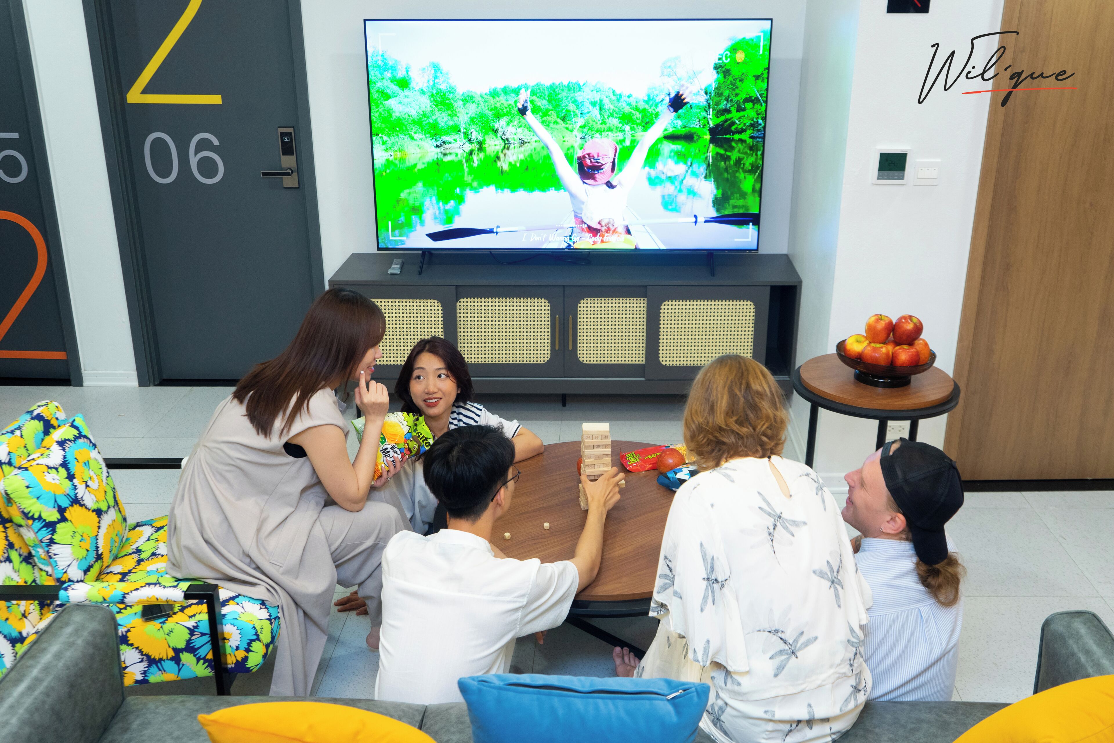 A group of people playing a board game in a modern living room with a television displaying a nature scene.
